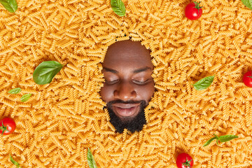 Overhead shot of dark skinned bearded man keeps eyes shut has thick beard buried in raw macaroni with red tomatoes and basil leaves. Ingredients for making pasta. Food and nutrition concept.