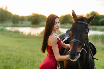 Obraz premium A young pretty girl in a red dress poses on a ranch with a thoroughbred stallion at sunset. Love and care for animals