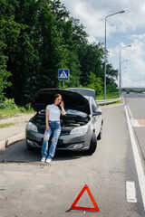 A young girl stands near a broken-down car in the middle of the highway and calls for help on the phone. Failure and breakdown of the car. Waiting for help.