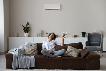 Peaceful African American millennial woman using air conditioner remote controller, relaxing on...