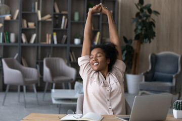 Happy carefree African American woman stretching hands at workplace, leaning back in comfortable...