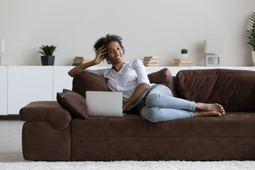 Full length portrait of smiling African American woman relaxing with laptop, lying on cozy couch at home, happy attractive young female looking at camera, enjoying leisure time with modern device