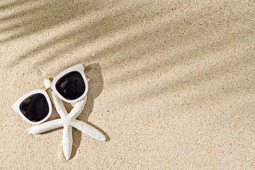 Fototapeta na wymiar Starfish and white sunglasses with palm leaf shadow on natural sand background, copy space