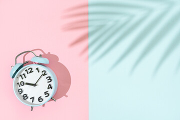 Old fashion blue alarm clock on pink and blue background with palm leaf shadiw, flat lay with copy space