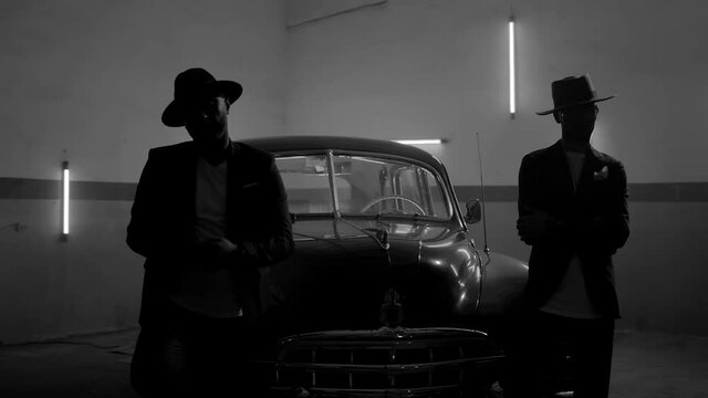 Two silhouettes of men in black suits with classic hats standing by vintage old car. Strangers look like italian mafioso from movie . Footage have permissible count of normally film effect style GRAIN