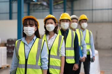 Group of Asian male and female engineers team wearing hygienic mask protect with helmet safety stand in line in factory warehouse