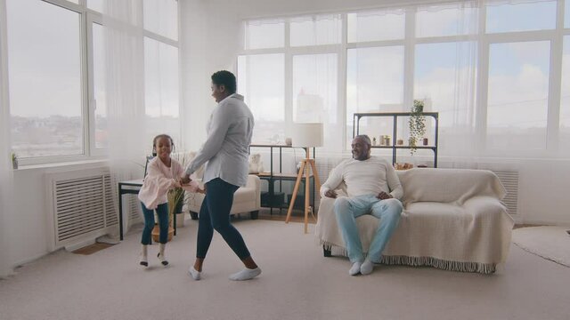 Afro American mother spins rotates little daughter and plays game with baby while black father sits on couch in living room. Loving daddy jumping into air with female child at home, family together