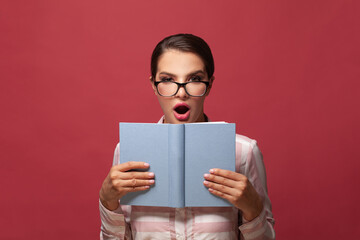 Portrait of smart surprised woman in glasses holding book with empty cover posing isolated on red wall background. Mock up copy space. People sincere emotions lifestyle concept