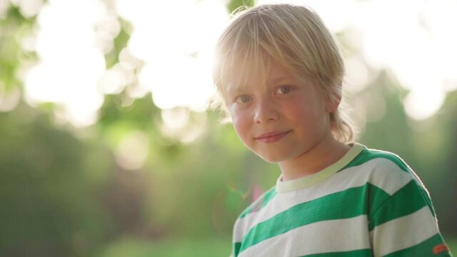Cute blonde little boy looking at camera in sunlight, copy space to left. Kid in striped t-shirt posing outside on sunny summer day, slow motion head and shoulders portrait