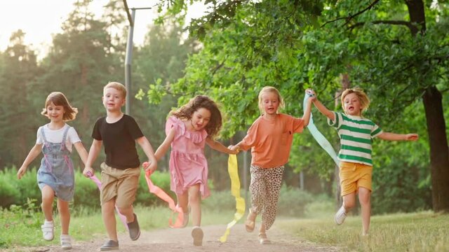 Group of five happy kids running on grass in park holding hands with colorful ribbons and screaming with excitement. Children enjoying freedom and togetherness, front view full length slow motion shot
