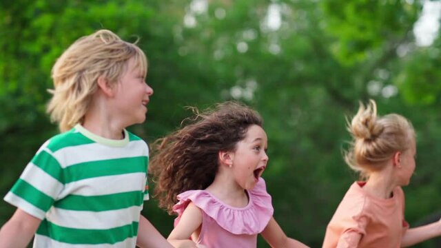 Group of happy kids running outdoors in green park holding hands and screaming with excitement. Playful children enjoying friendship and togetherness, side view slow motion medium shot