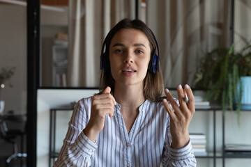 Young business woman in headphones making video call, looking at camera, speaking at webcam, having...