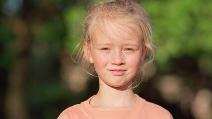 Closeup head and shoulders portrait of cute blonde little girl looking at camera standing outside in park on windy summer day. Kid smiling slightly in wind