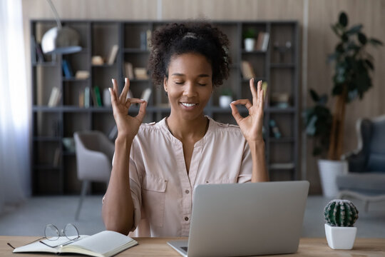 Smiling African American young woman with closed eyes meditating, sitting at desk with laptop, mindful happy businesswoman freelancer or student enjoying break, practicing yoga, stress relief concept