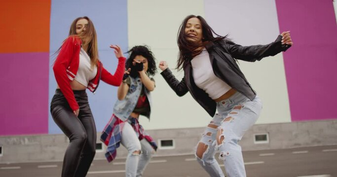 4K Three young beautiful , happy girls dancing , moving , looking at camera . Stylish bloggers in sport outfit  having fun , jumping and shaking body in front of colorful background wall . slow motion
