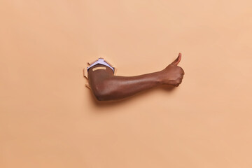 Picture of a muscled dark skinned hand, breaking through the paper background, showing thumb up. Has the plaster on the shoulder. Shows motivation for vaccination. Isolated on the beige background.