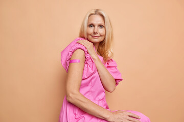 Middle aged smiling beautiful long haired blond woman sitting in a summer pink dress, shows her shoulder with pink band aid after getting the vaccine. Healthcare concept. Isolated over beige. 
