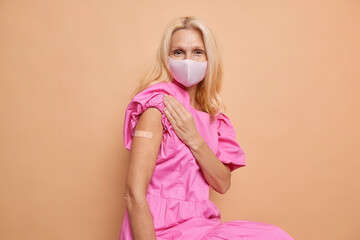 Nice serious caucasian grown up lady in a pink dress, wears a face mask. Holds her volumetric sleeve to show her shoulder with beige plaster on it after anticovid vaccination. Isolated over beige.