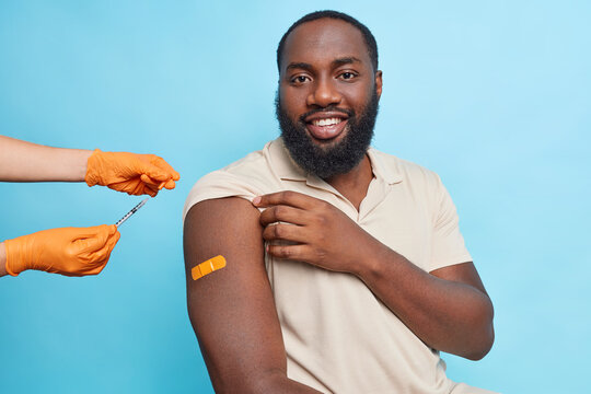 Muscled smiling ethnic man with a beard breathes out after vaccination and shows his orange plaster on the shoulder. Hands in orange gloves cover syringe with a cap. Isolated over blue background.
