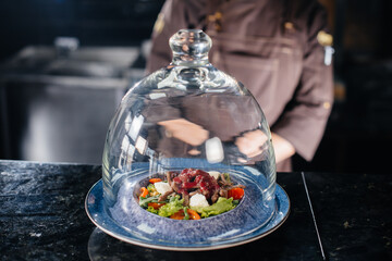 A professional chef serves a freshly prepared salad of tomato and veal greens with sauce in a refined restaurant under a glass dome.