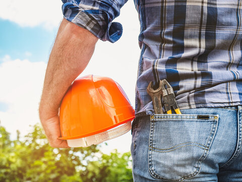 Attractive man in work clothes, holding tools in his hands against the background of trees, blue sky and sunset. View from the back. Labor and employment concept