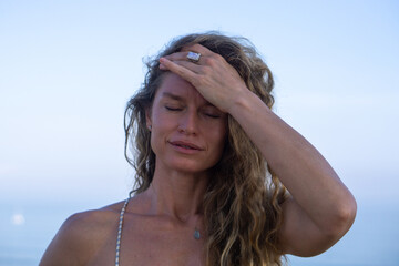Young woman with curly long blonde hair and blue eyes touching forehead with a hand  on cliff with ocean view and blue sky on background