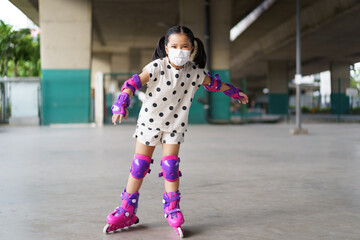 Asian child cute or kid girl exercise playing pink rollerblade or inline skates on sport skating rinks or park and wearing knee to wrist support and elbow pads with white face mask for safety protect