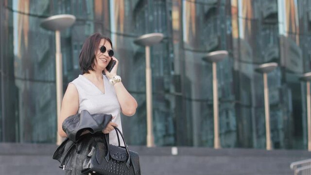 A businesswoman in round sunglasses stopped on the street to answer an incoming call on a cell phone. She talks emotionally with someone and looks in different directions.