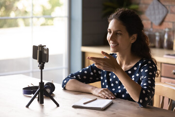 Happy Hispanic blogger girl taking video on mobile phone with tripod, speaking at webcam. Employee,...