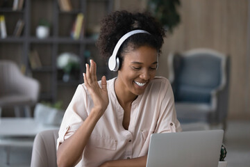 Close up smiling African American woman in headphones waving hand, using webcam, looking at laptop...