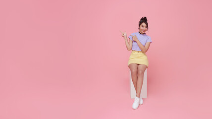 Obraz na płótnie Canvas Happy beautiful Asian teen woman sitting on chair and hands pointing on pink background with copy space. panoramic background.