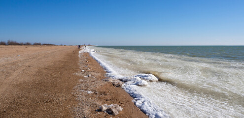 Winter beach of the sea with frozen sand and ice blocks and snow in the water. Snow-icy Cold dunes along the seashore. In the distance, people are walking along the coastline. Winter seascape