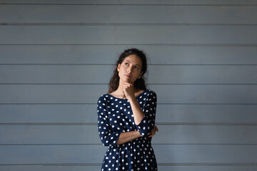 Thoughtful millennial Hispanic woman in spotted dress touching chin in deep thoughts, looking away, thinking of choice, offer, opportunities, future success, making decision. Head shot portrait