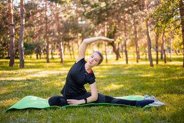 Outdoor Pilates training. A young woman does exercises to stretch the muscles of her legs while sitting on a yoga mat in the park. Exercise for the development of twine.