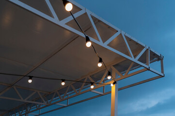 Textile visor on canopy with garland of energy saving lamps illuminating terrace of restaurant of night city