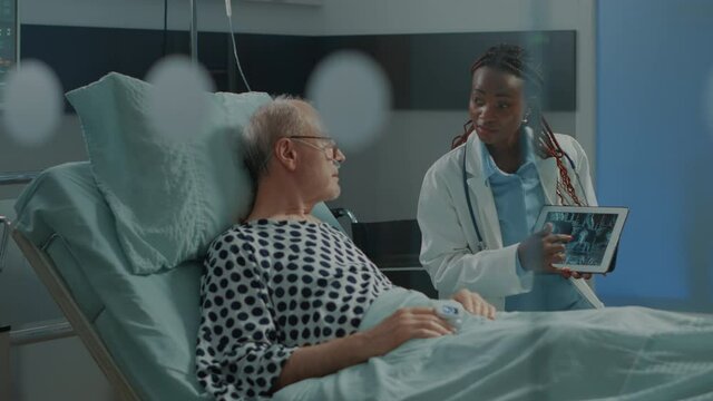 African american medic explaining xray on tablet to patient in hospital ward bed at medical facility. Sick old man learning about health problems and lung diagnosis to start treatment