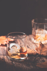 Glass of scotch whiskey and ice on wooden background with autumn leaves