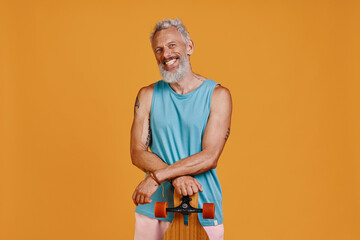 Carefree senior man carrying skateboard and smiling while standing against orange background
