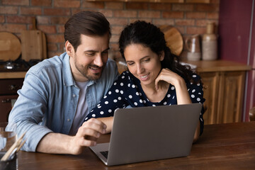 Happy millennial family couple surfing internet on laptop, shopping online together, making order, booking hotel, using app or service. Man and woman pointing at computer screen in kitchen at home