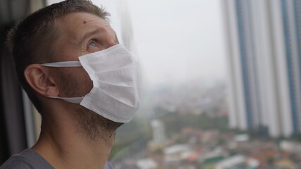 Face of a man in face mask standing by the window with his eyes closed as a sign of exhaustion of...