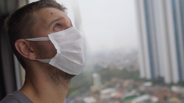 Face of a man in face mask standing by the window with his eyes closed as a sign of exhaustion of the isolation