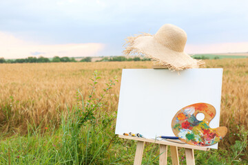 Wooden easel with blank canvas, painting equipment and hat in field. Space for text