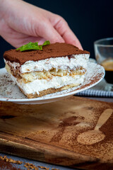 woman taking traditional tiramisu cake on a plate. Authentic gluten-free Italian layered dessert with ladyfinger biscuits, mascarpone cheese cream and cocoa powder. Cup of coffee in the background.