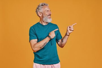 Happy senior man pointing copy space and smiling while standing against orange background