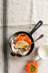 Dutch baby pancake with fresh persimmon and banana with icing sugar powder in red pan on white kitchen background. Top view