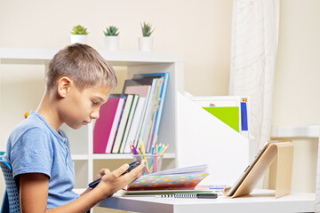 Teenage boy sitting at the table with ldigital tablet computer, notebooks and scrolling phone instead of doing homework at home. Technology, online learning, entertainment at home