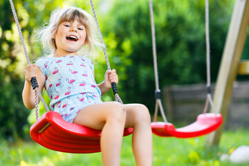 Happy little toddler girl having fun on swing in domestic garden. Smiling positive healthy child swinging on sunny day. Preschool girl laughing and crying. Active leisure and activity outdoors. - obrazy, fototapety, plakaty