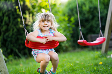 Happy little toddler girl having fun on swing in domestic garden. Smiling positive healthy child...