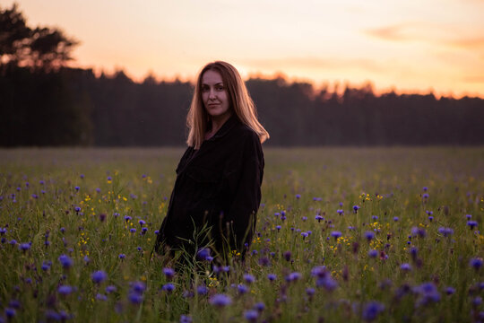 Young woman in a black jacket walks in a cornflower field and looks at camera at sunset. Amazing summer landscape in Belarus. Beautiful orange sunset over blue wildflowers meadow.