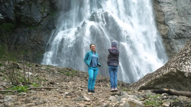 Cute Little Girl Taking Photos of her Mother by Beautiful Waterfall with the Smartphone, Slow Motion. Ecotourism and Travel Lifestyle Concept